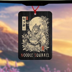 Może przedstawiać: Czarny drewniany odświeżacz powietrza z japońską ilustracją wiewiórki jedzącej makaron. Projekt zawiera bambus, pełnię księżyca i japoński tekst. Słowa "NOODLE SQUIRREL" są wydrukowane na czerwono.