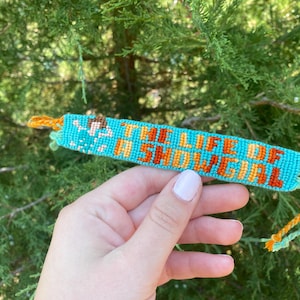 Puede incluir: Una pulsera de la amistad turquesa con el texto "THE LIFE OF A SNOWGIRL" en naranja. La pulsera tiene un dise&ntilde;o tejido con hilos naranjas y amarillos en los extremos. El fondo es una planta verde y frondosa.