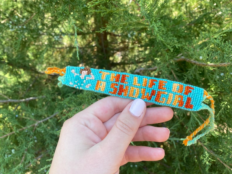 Puede incluir: Una pulsera de la amistad turquesa con el texto "THE LIFE OF A SNOWGIRL" en naranja. La pulsera tiene un dise&ntilde;o tejido con hilos naranjas y amarillos en los extremos. El fondo es una planta verde y frondosa.