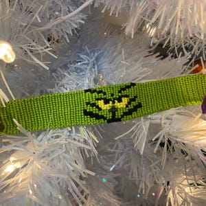 May include: Green and black friendship bracelet with a green and yellow Grinch face design.