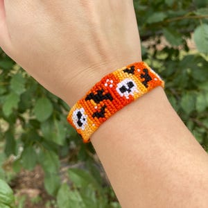 May include: A handmade friendship bracelet in orange, yellow, black, and white. The woven bracelet features a repeating pattern of skulls and geometric shapes. The bracelet is worn on a wrist against a background of green foliage.
