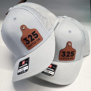 Puede incluir: Dos gorras de camionero gris claro con la parte trasera de malla blanca. Cada gorra presenta un parche de cuero marrón con el número "325" y "CATTLE CO." impreso en negro. Las gorras tienen una visera curva y una correa ajustable.