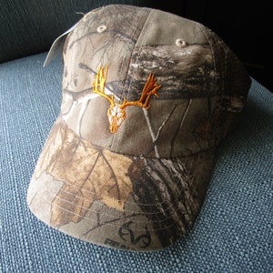 May include: Camouflage baseball cap with an embroidered orange deer skull design.