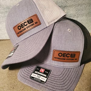 May include: Two gray and white trucker hats with a brown leather patch that reads "OEC Petroleum Systems Inc.". The hats have a black mesh back and a black adjustable strap with a red and white label that reads "Est. 1970 Richardson Original 112 Trucker Adjustable".