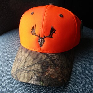 May include: A bright orange baseball cap with a camouflage brim. The cap has a black embroidered deer skull with antlers on the front.