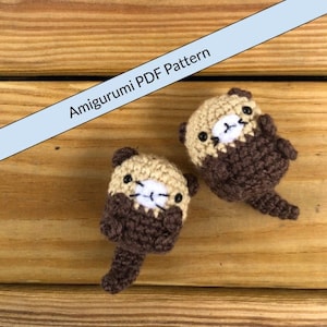 以下が含まれることがあります： 2つの編みぐるみカワウソのぬいぐるみ。1つのカワウソは茶色で顔はベージュ、もう1つはベージュで顔は茶色。両方のカワウソは黒い目と白い鼻をしています。カワウソは木製の表面に座っています。テキスト「Amigurumi PDF Pattern」は青い背景に書かれています。
