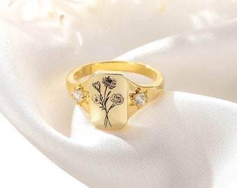 Puede incluir: Un anillo dorado con una cara rectangular con un diseño floral negro. El anillo tiene dos gemas redondas y transparentes a cada lado de la cara. El anillo está sobre un fondo blanco y sedoso.