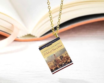 May include: A gold-colored chain necklace with a miniature book pendant. The book cover features the title "A Tale of Two Cities" by Charles Dickens. The background includes a blurred open book.