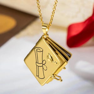 May include: A gold-colored graduation-themed pendant necklace. The pendant is a square locket with a scroll and mortarboard design. The necklace has a delicate chain and a clasp to open the locket. The background is blurred with a red ribbon.