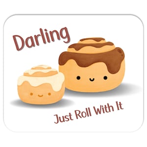 May include: A white background with the text "Darling" in brown lettering. Two cartoon cinnamon rolls with smiling faces. The larger cinnamon roll has chocolate icing and the smaller cinnamon roll has white icing. The text "Just Roll With It" is in brown lettering below the cinnamon rolls.