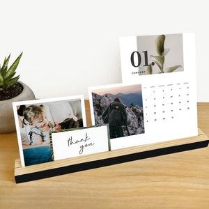 以下が含まれることがあります： 黒い縁取りのある木製デスクトップカレンダーホルダー。ホルダーには3枚のカードが表示されています。1月はカレンダーカード、ハイキングをしている人の写真カード、子供と猫の写真カードです。カレンダーカードには「01. 1月」というテキストが、写真カードには「ありがとう」というテキストが書かれています。