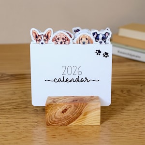 May include: A white 2026 calendar with a wooden base. The calendar features illustrations of four different dog breeds peeking over the top. Black paw prints are on the right side of the calendar.