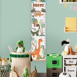 May include: A vertical hanging growth chart featuring a white background with colorful dinosaur illustrations, tropical plants, and a volcano. The name BRETT is printed in brown letters. The chart includes measurement scales in both inches and centimeters.