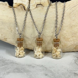May include: Three silver chain necklaces with small glass bottles filled with white bone fragments. The bottles have cork stoppers and are hanging from the chains.