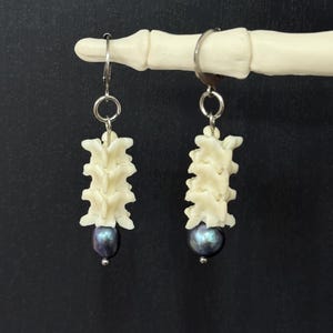 May include: A pair of drop earrings featuring a stack of three off-white, bone-like vertebrae segments suspended from a silver-toned metal hoop, finished with a single dark, iridescent, round bead at the bottom of each earring structure.