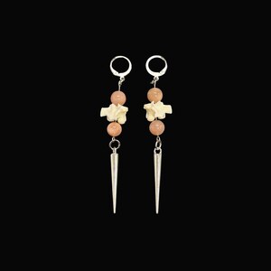 May include: A pair of dangle earrings with a unique design. Each earring features a silver-colored spike pendant, two peach-colored beads, and a bone-shaped charm. The earrings hang from silver-colored ear wires against a black background.