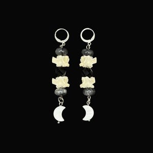 May include: A pair of dangle earrings featuring silver-tone leverback hooks, grey round beads, off-white textured bone-like segments, faceted black beads, and white crescent moon charms. The earrings are arranged vertically with alternating beads.