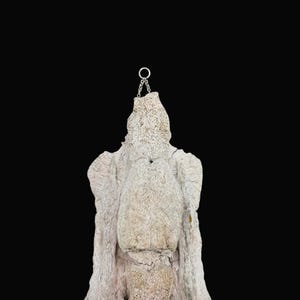 May include: A textured, off-white sculpture with a chain and ring at the top, set against a black background. The sculpture has a rough, organic appearance, with visible ridges and a layered texture. The chain and ring suggest it may be designed for hanging.