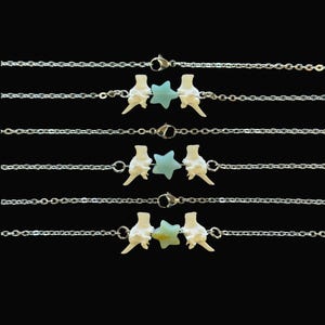 May include: Several silver chain bracelets, each featuring a bone-like charm flanked by a light blue star-shaped bead. The bracelets are displayed against a black background, showcasing the delicate design and craftsmanship of the jewelry.