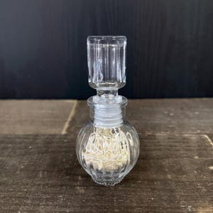 May include: A clear, small glass perfume bottle with a ribbed, round body and a rectangular stopper. The bottle has a white stopper and is displayed on a wooden surface against a dark background.