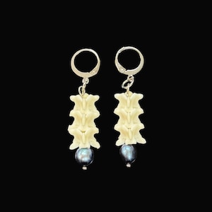 May include: A pair of drop earrings featuring silver-toned leverback hooks, each suspending a vertical stack of three off-white, bone-like vertebrae segments, finished with a single dark, iridescent, round pearl at the bottom of each earring.