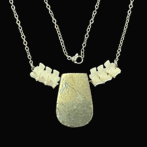 May include: A unique necklace featuring a silver chain with a pendant of a gold-colored, teardrop-shaped stone. The necklace also includes two clusters of small, off-white bone-like pieces on either side of the pendant. The background is black.