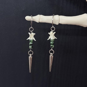 May include: A pair of dangle earrings featuring a white bone vertebra segment, three small green speckled beads, and a silver-toned metal spike charm suspended from a silver-toned metal ear wire hook. The design is symmetrical and linear.