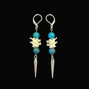 May include: A pair of dangle earrings with a unique design. Each earring features a silver-colored spike pendant, teal and blue beads, and a bone-shaped element. The earrings hang from silver-colored lever-back closures against a black background.