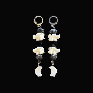 May include: A pair of dangle earrings with a celestial theme. Each earring features a gold or silver hoop, dark gray beads, white bone-shaped charms, black beads, and a white crescent moon charm. The earrings are displayed against a black background.