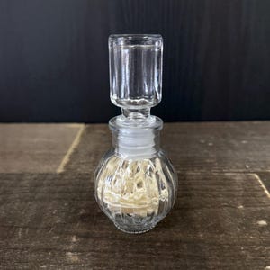 May include: A clear glass perfume bottle with a ribbed body and a cylindrical stopper. The bottle contains a light-colored material and sits on a wooden surface. The background is a dark, solid color.