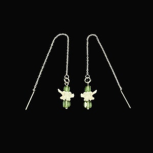 May include: A pair of silver-toned chain threader earrings featuring a central off-white bone-shaped charm flanked by two faceted translucent green beads on each earring. The delicate metal chains hang vertically from the central decorative element.