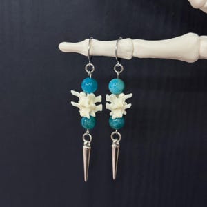 May include: A pair of dangle earrings featuring silver-toned metal hooks, each holding a blue spherical bead, a small off-white bone-like vertebra segment, another blue spherical bead, and a pointed silver-toned metal spike at the bottom.