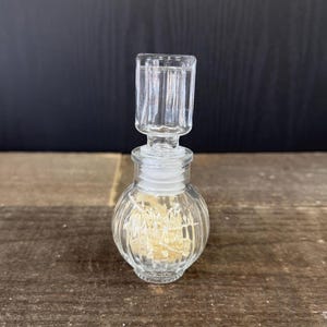 May include: A clear, ribbed glass perfume bottle with a matching stopper. The bottle has a round body and a cylindrical neck, with a clear stopper. The bottle is sitting on a wooden surface.