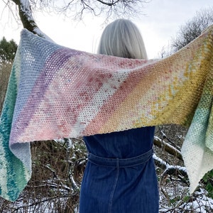 以下が含まれることがあります： レース編みの虹色のグラデーションのショール。ショールは人の肩にかけられており、風になびいている。