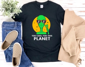 May include: Black t-shirt with a green alien wearing sunglasses and making a peace sign. The text on the shirt reads "Too Cool For This Planet".