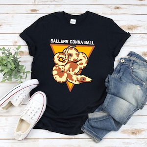 Puede incluir: Camiseta negra con un triángulo amarillo y una serpiente marrón y blanca. El texto "Ballers Gonna Ball" está impreso en la camiseta.