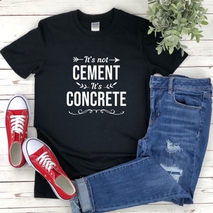 May include: Black t-shirt with white text that reads "It's not cement, It's concrete" with a decorative arrow and floral design. The shirt is displayed on a wooden surface with a pair of red sneakers and blue jeans.