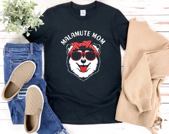 Pode incluir: Camiseta preta com um cão Malamute branco usando óculos de sol vermelhos e uma bandana vermelha com bolinhas brancas. O texto "MALAMUTE MOM" é impresso em branco acima do cão.