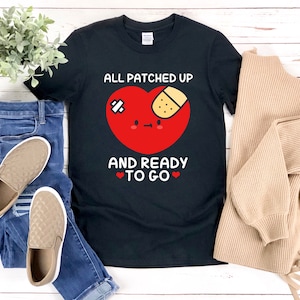 Op de afbeelding: Zwart T-shirt met een rode hartgrafiek en de tekst "All patched up and ready to go". Het hart heeft een pleister en een schattig gezicht.