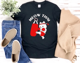 May include: Black t-shirt with the words "MEOW THAI" above a cartoon cat kicking a red punching bag. The cat is wearing a red headband, boxing gloves, and shorts. The shirt is displayed with blue jeans, tan shoes, and a tan sweater.