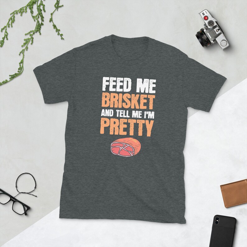 May include: A dark heather grey t-shirt with the text "Feed me brisket and tell me I'm pretty" in white. A cartoon image of a slice of brisket is below the text.