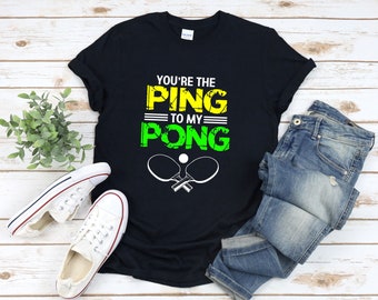 Könnte beinhalten: Schwarzes T-Shirt mit einer grünen und gelben Grafik, auf der "You're the Ping to my Pong" steht, mit zwei gekreuzten Tischtennis Schlägern.