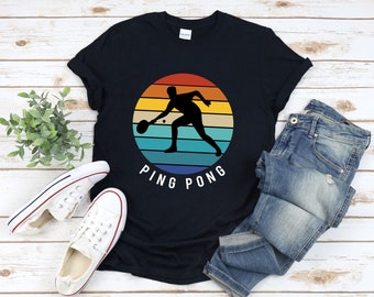 Könnte beinhalten: Ein schwarzes T-Shirt mit einer Retro-Grafik einer Person, die Tischtennis spielt. Die Grafik ist in einem Kreis mit einem Regenbogen-Farbschema. Der Text "PING PONG" befindet sich unter der Grafik.