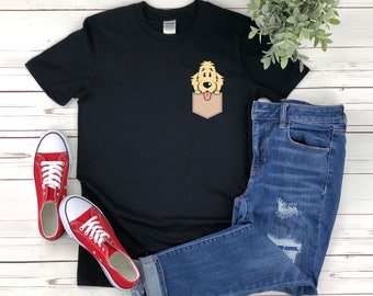 Puede incluir: Una camiseta negra con un gráfico de un perro golden retriever asomándose de un bolsillo. La camiseta está colocada sobre una superficie de madera blanca con un par de zapatillas rojas y unos vaqueros azules.