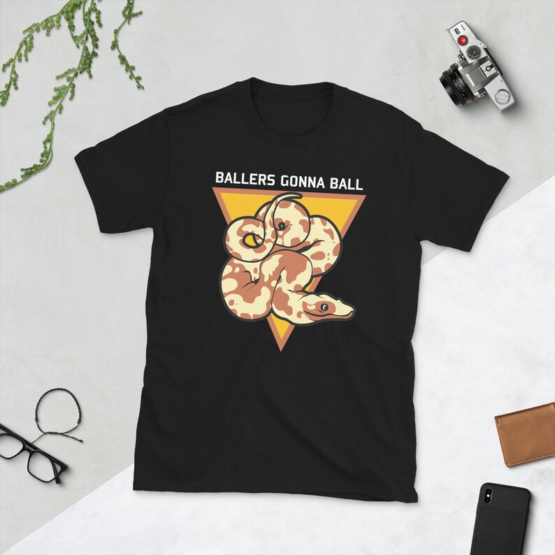 Op de afbeelding: Zwart T-shirt met een witte driehoek en de tekst "Ballers Gonna Ball" in wit. Binnen de driehoek is een cartoonillustratie van een bruine en witte slang.