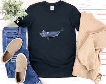 May include: Black t-shirt featuring a graphic of a cat swimming like a fish. The cat is grey and has a white belly. The t-shirt is on a white wooden surface with a pair of blue jeans and brown shoes.