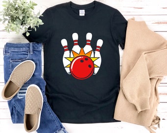 Pode incluir: Camiseta preta com um gráfico de pinos de boliche e uma bola de boliche. Os pinos de boliche são brancos com detalhes vermelhos e amarelos. A bola de boliche é vermelha com pontos brancos.