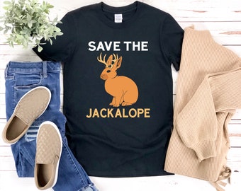 May include: Black t-shirt with the text "SAVE THE JACKALOPE" above and below a cartoon jackalope illustration. The jackalope is brown with antlers. The shirt is displayed with blue jeans, tan slip-on shoes, and a tan sweater.