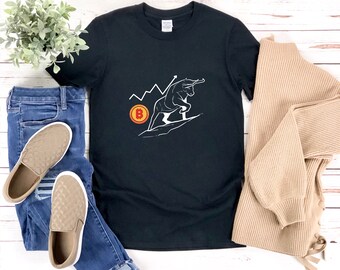 May include: Black t-shirt with a white graphic of a bull charging up a line chart with a Bitcoin symbol. The shirt is laid out on a white wooden surface with a pair of brown quilted slip-on shoes and blue jeans.