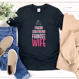 Puede incluir: Camiseta negra con texto blanco que dice "Friend, Girlfriend, Fianc&eacute;e" con una l&iacute;nea a trav&eacute;s de cada palabra. La palabra "Wife" est&aacute; en rosa debajo de las otras palabras. La camiseta est&aacute; sobre una superficie de madera blanca con un par de zapatos acolchados marrones y unos vaqueros azules.
